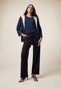 Vrouw in navy blauwe broek, bijpassende top en een tweekleurige blazer, poserend met één hand in haar zak en zwarte hoge hakken.