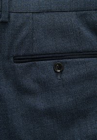 Nahaufnahme eines dunkelblauben, strukturierten Stoffes, der eine Gesäßtasche mit schwarzem Knopf und horizontalem Paspelöffnungsdesign an maßgeschneiderten Hosen zeigt.