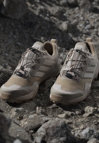 Beżowe buty outdoorowe z teksturowaną cholewką, gumową podeszwą z gripem i elastycznymi sznurowadłami. Posiadają trzy ukośne paski po każdej stronie.