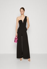 Svart en-skulder jumpsuit med dyp V-hals, vide ben og lommer. En blank rosa håndtaske og gjennomskinnelige, utsmykkede hæler fullfører antrekket.