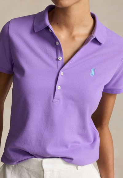 Γυναίκα που φοράει κοντομάνικο πολο σε ανοιχτό μοβ χρώμα με λευκά κουμπιά και μικρό κεντημένο λογότυπο σε τυρκουάζ στο στήθος.