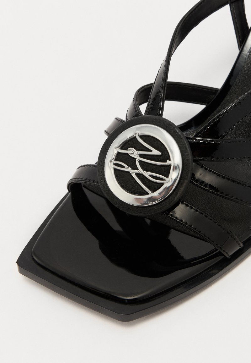 Sandale en cuir verni noir avec des brides croisées. Présente un accent métallique rond au centre et un design de bout carré.