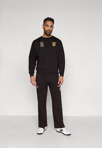 Felpa nera e pantaloni abbinati con dettagli di marca in oro e rosso. Misto cotone testurizzato, scollo rotondo, vestibilità comoda e sneakers bianche.