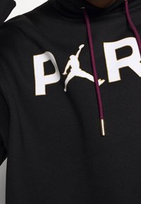 Sweat à capuche noir avec un grand "P" blanc et un accent doré, accompagné du logo Jumpman. Cordon violet avec embouts dorés. Texture de tissu douce.