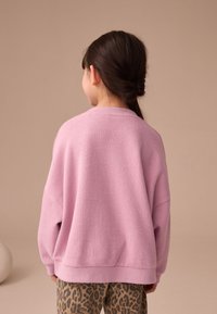 Svetloružový nadrozmerný sveter z mäkkej látky s rebrovaným výstrihom a manžetami, skombinovaný s hnedými nohavicami s leopardím vzorom.