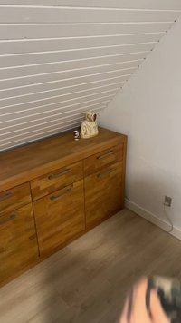 Medinė spintelė su metalinėmis rankenomis po kreiva balta lentjuosčių lubomis, ant jos stovi maža figūrėlė ir skulptūra su gobtuvu.