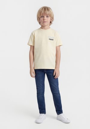 Γάλλος αγόρι στέκεται μπροστά, φοράει ανοιχτόκίτρινη μπλούζα με κείμενο, μπλε τζιν και λευκά αθλητικά παπούτσια σε λευκό φόντο.