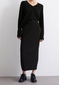 Schwarzer Strickpullover mit V-Ausschnitt, kombiniert mit einem schwarzen, gerippten Midirock. Grobe schwarze Schnürschuhe vervollständigen das Outfit.