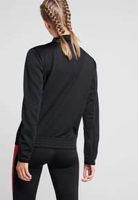 Schwarze Sportjacke mit glatter Textur, ausgestattet mit einem Reißverschlusskragen und gerippten Bündchen. Rote Leopardenmusterakzente an den Seiten.