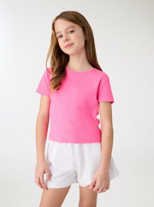 Ragazza con capelli lunghi che indossa una maglietta rosa acceso a maniche corte e pantaloncini bianchi, in piedi davanti a uno sfondo chiaro e uniforme.