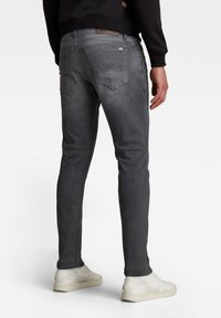 Homme portant un jean noir délavé slim et des baskets blanches, debout de dos, devant un fond clair uni.