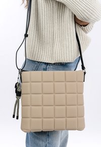 Borsa a tracolla beige trapuntata con design imbottito, tracolla nera regolabile e dettagli decorativi con frange in bianco e nero.