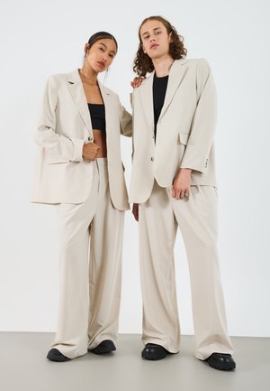 Blazer oversized beige con ampi revers su top neri, abbinato a pantaloni beige larghi e scarpe nere chunky, su uno sfondo neutro.