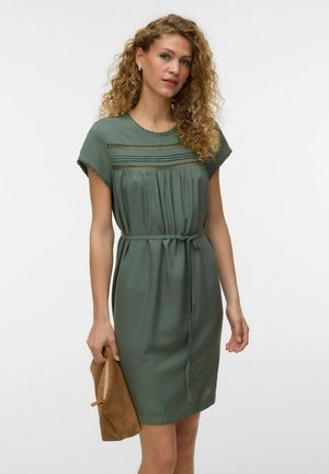 Femme aux cheveux bouclés portant une robe verte ceinturée avec des incrustations en dentelle, tenant un chapeau en daim marron dans sa main gauche sur un fond uni.