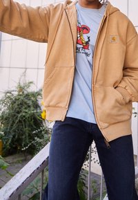 Sudadera con cremallera color caqui y un bolsillo frontal, llevada sobre una camiseta gráfica color azul claro; cuenta con un corte relajado y puños acanalados.