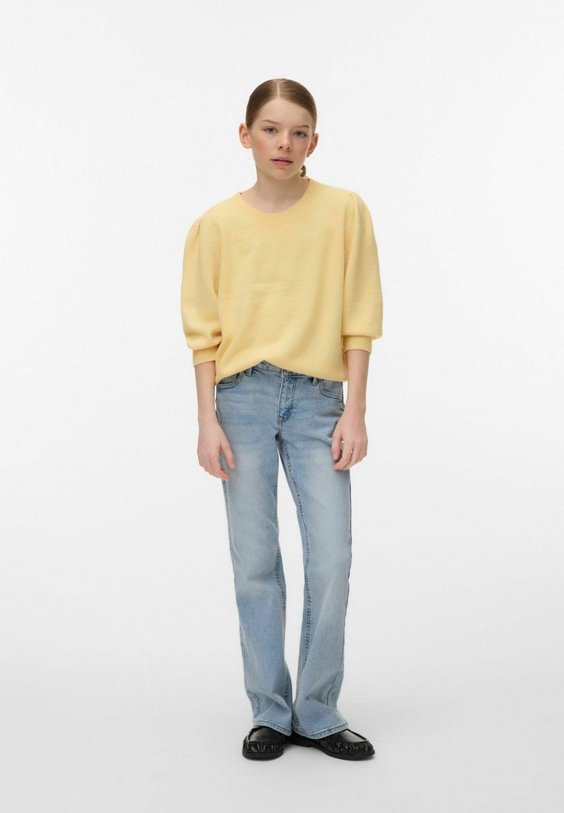 Jeune personne debout portant un pull jaune pâle, un jean droit bleu clair et des chaussures noires, sur un fond blanc uni.