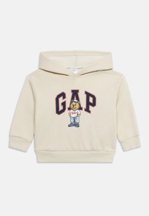 Kremowa dziecięca bluza z kapturem z logo GAP oraz kreskówkowy miś ubrany w dżinsy, białą koszulkę GAP i czapkę z daszkiem z przodu.