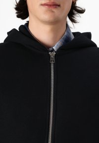 Schwarze Zip-Hoodie mit einem Metallreißverschluss, weicher Textur und einer Kapuze. Darunter ist teilweise ein blau kariertes Hemd sichtbar.