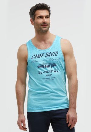 Man wearing light blue sleeveless shirt with text "Camp David" and dark pants, standing against a plain light background.