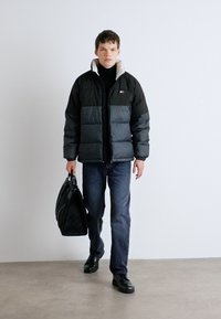Giacca puffer nera e grigia, con colletto alto e design trapuntato. Abbinata a jeans scuri e scarpe nere. Tiene una borsa nera.