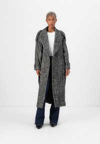 Cappotto lungo e oversize in pattern houndstooth, realizzato in una miscela di lana grigia e nera, con ampi rever e vita chiusa da una cintura.