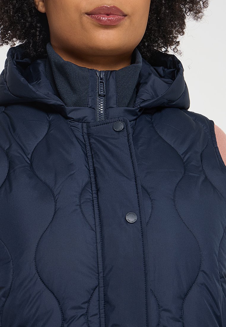 Gilet matelassé bleu marine avec capuche, doté d'une fermeture éclair et de boutons-pression ; tissu texturé avec un motif ondulé et un col côtelé.