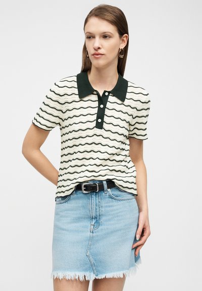 Γυναίκα που φοράει μια κοντομάνικη πόλο μπλούζα με άσπρες και μαύρες κυματιστές ρίγες, με μαύρο γιακά, και μια ανοιχτό μπλε φθαρμένη τζιν φούστα.