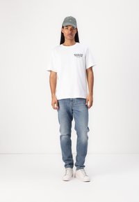 White cotton t-shirt with "REPUBLIQUE FRANCAISE" text, light blue jeans, and light grey baseball cap. White sneakers complete the outfit.