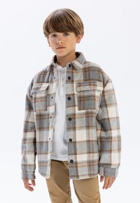 Chemise en flanelle à carreaux gris, blanc et marron avec deux poches avant et boutons noirs, portée sur un henley blanc. Un pantalon marron complète la tenue.
