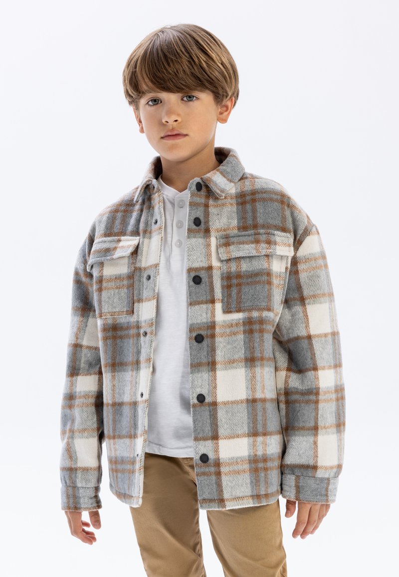 Chemise en flanelle à carreaux gris, blanc et marron avec deux poches avant et boutons noirs, portée sur un henley blanc. Un pantalon marron complète la tenue.