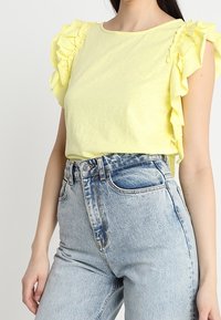 Žlutý krátký rukávový top s volánky, kombinovaný s vysokými světle modrými džíny. Tkanina vypadá měkce a má volný střih.