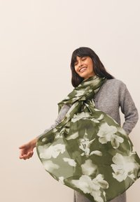Sciarpa verde con motivo floreale, realizzata in tessuto leggero e testurizzato, che cade morbida sulle spalle, accentuando un look a strati.