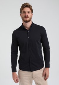 Camisa negra de manga larga con botones, hecha de una tela ajustada y con cuello. Llevada con pantalones de color claro.