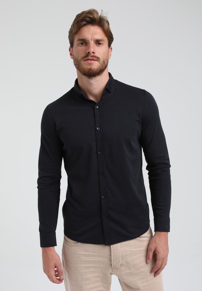 Camisa negra de manga larga con botones, hecha de una tela ajustada y con cuello. Llevada con pantalones de color claro.