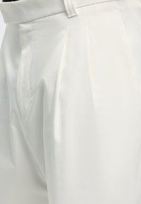Pantalon blanc sur mesure avec plis, ceinture et passants, présenté en gros plan sur la texture et les coutures du tissu.