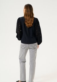 Chemise noire avec manches bouffantes en dentelle, associée à un jean gris clair à jambes droites. Le tissu semble texturé avec des détails en dentelle complexes.