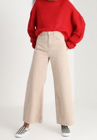 Jeans beige a gamba larga con vita alta, abbinati a un maglione rosso di lana spesso e scarpe slip-on a quadri in bianco e nero.