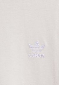 Bílá tkanina s malým vyšitým logem Adidas ve tvaru trojlístku a názvem značky ve světle šedé nitě.