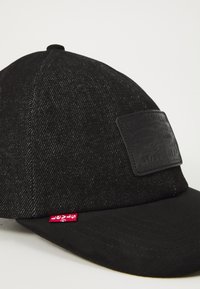 Zwarte pet met een denimstructuur, voorzien van een leren patch aan de voorkant, een gebogen klep en een klein rood merklabel aan de zijkant.