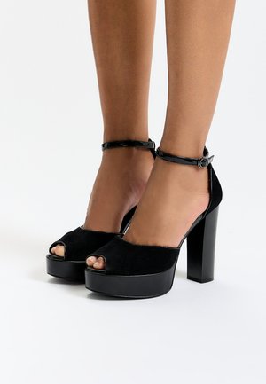 Sorte peep-toe platform hæle med en overdel i ruskind, akkompagneret af en patent ankelrem og en chunky hæl, der har en glat overflade og let glans.