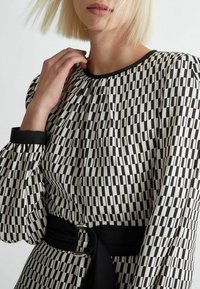 Donna con un bob biondo che indossa un vestito a maniche lunghe con un motivo geometrico bianco e nero e una larga cintura nera, con la mano vicino alla clavicola.