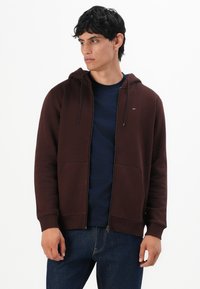 Joven con una sudadera con cremallera marrón oscuro sobre una camiseta azul marino y jeans oscuros, mirando ligeramente hacia la izquierda contra un fondo blanco.