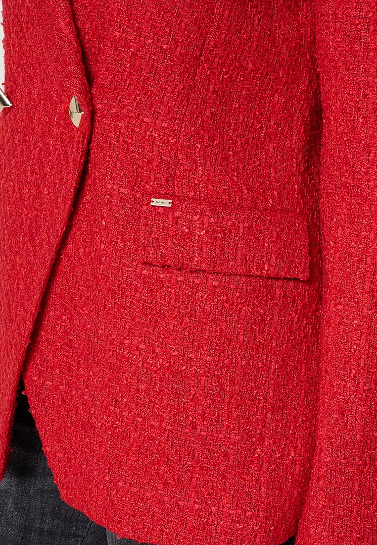 Blazer rosso texturizzato con rever a punta, tasca singola e piccolo logo metallico. Il materiale sembra tessuto e vivace.