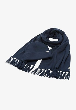 Sciarpa a coste blu navy con bordi sfrangiati, abbinata a un berretto coordinato. Entrambi i pezzi presentano un dettaglio etichetta sottile. Texture morbida.