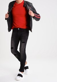 Hombre con camiseta roja, chaqueta de cuero negra con rayas rojas, jeans rotos negros y zapatillas blancas sin cordones con tiras negras.