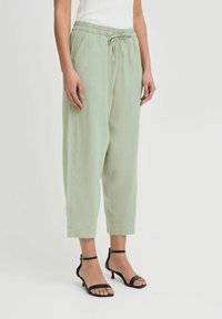 Pantalons verts clairs, amples avec une taille élastique et un cordon de serrage. Comprend des poches latérales et des poignets fuselés. Portés avec des talons ouverts noirs.