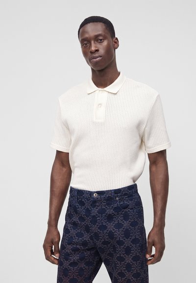 Homme portant un polo en maille blanc à manches courtes et un pantalon bleu foncé à motifs, debout devant un fond gris clair uni.