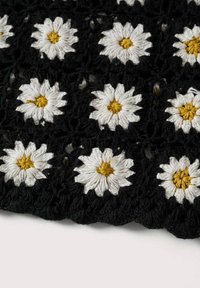 Zwarte gehaakte stof met witte en gele geborduurde madeliefjes in een rasterpatroon, met een opvallende textuur en ingewikkeld ontwerp.