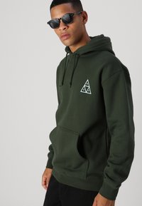 Sudadera verde con bolsillo tipo canguro, capucha ajustable y logo blanco en forma de triángulo. Textura suave, corte casual, combinada con gafas de sol negras.