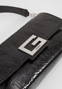 Pochette Guess en cuir verni noir avec motif de logo embossé et grande fermeture métallique argentée en forme de G.